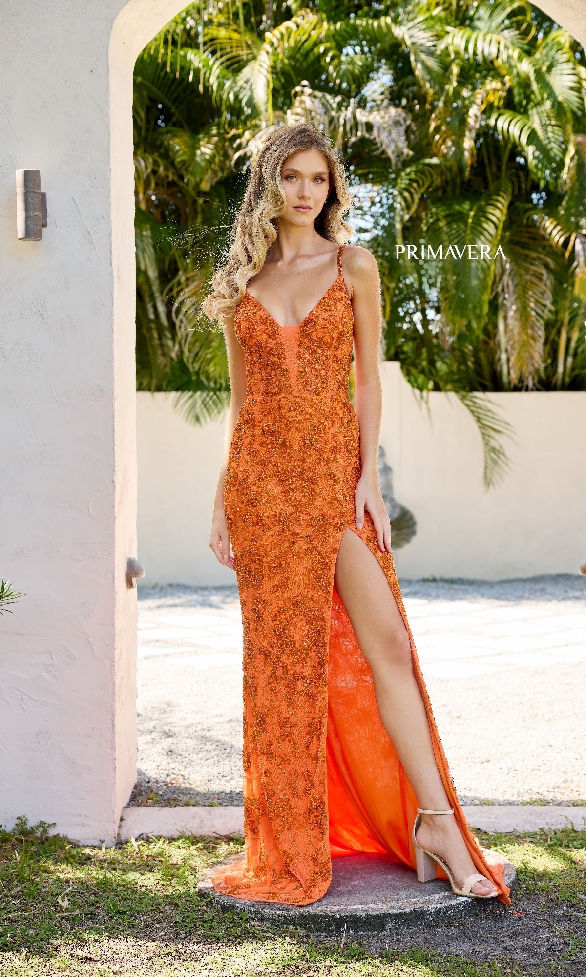 A woman poses near a white archway with greenery, wearing the Primavera 4317 Lace-Up Long Lace Prom Dress in bright orange, featuring embroidered lace, spaghetti straps, and a thigh-high slit.